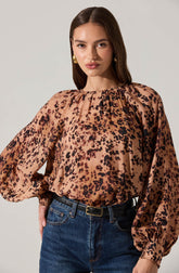 A woman is wearing a long-sleeved blouse with a leopard print pattern in shades of tan, brown, and black. The blouse has a gathered neckline with subtle pleats and voluminous sleeves that are cinched at the wrists. She is also wearing high-waisted blue jeans with a black belt.